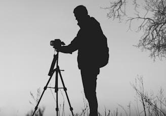 silhouette photo of man in front of DSLR camera with tripod under leafless tree