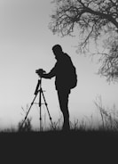silhouette photo of man in front of DSLR camera with tripod under leafless tree