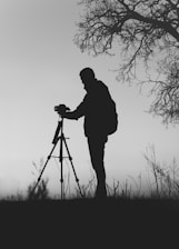 silhouette photo of man in front of DSLR camera with tripod under leafless tree