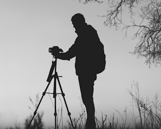 silhouette photo of man in front of DSLR camera with tripod under leafless tree
