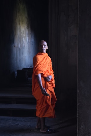 A person wearing a bright orange robe stands in a dimly lit, ancient-looking architectural setting. The light casts subtle shadows on the stone walls, creating a serene and contemplative atmosphere.