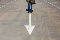 A person stands on a large, white arrow painted on a concrete surface. The individual is wearing jeans and brown shoes, with a bag slung over their shoulder and a camera hanging by their side. The scene suggests a sense of direction or choice, with an expansive, empty space around them.