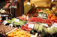 A market stall displays a vibrant variety of fresh fruits and vegetables. Trays of asparagus, tomatoes, oranges, lettuce, and walnuts are arranged neatly. The produce is labeled with price tags, and the entire scene is well-lit with warm lighting.