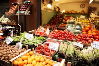A market stall displays a vibrant variety of fresh fruits and vegetables. Trays of asparagus, tomatoes, oranges, lettuce, and walnuts are arranged neatly. The produce is labeled with price tags, and the entire scene is well-lit with warm lighting.