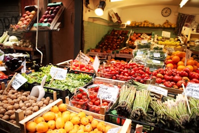 A market stall displays a vibrant variety of fresh fruits and vegetables. Trays of asparagus, tomatoes, oranges, lettuce, and walnuts are arranged neatly. The produce is labeled with price tags, and the entire scene is well-lit with warm lighting.