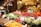 A market stall displays a vibrant variety of fresh fruits and vegetables. Trays of asparagus, tomatoes, oranges, lettuce, and walnuts are arranged neatly. The produce is labeled with price tags, and the entire scene is well-lit with warm lighting.