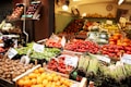 A market stall displays a vibrant variety of fresh fruits and vegetables. Trays of asparagus, tomatoes, oranges, lettuce, and walnuts are arranged neatly. The produce is labeled with price tags, and the entire scene is well-lit with warm lighting.