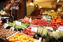 A market stall displays a vibrant variety of fresh fruits and vegetables. Trays of asparagus, tomatoes, oranges, lettuce, and walnuts are arranged neatly. The produce is labeled with price tags, and the entire scene is well-lit with warm lighting.