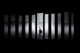 A shadowy figure moves behind frosted glass panels, creating a sense of mystery and ambiguity. The figure appears to be walking, distorted by the translucent glass that stretches vertically across the scene, casting an eerie atmosphere.