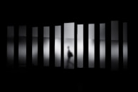 A shadowy figure moves behind frosted glass panels, creating a sense of mystery and ambiguity. The figure appears to be walking, distorted by the translucent glass that stretches vertically across the scene, casting an eerie atmosphere.