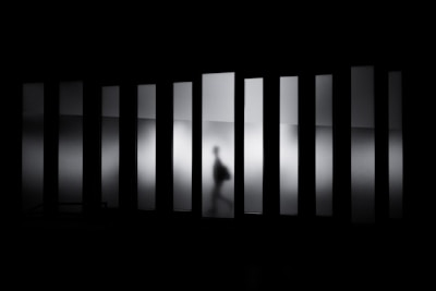 A shadowy figure moves behind frosted glass panels, creating a sense of mystery and ambiguity. The figure appears to be walking, distorted by the translucent glass that stretches vertically across the scene, casting an eerie atmosphere.