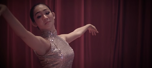 A dancer gracefully extends her arms while wearing a sleeveless sequined dress, performing against a backdrop of rich maroon curtains.