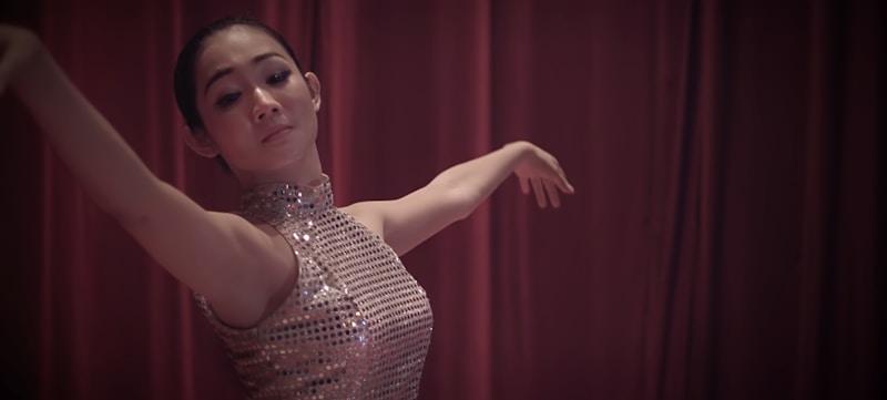 A dancer gracefully extends her arms while wearing a sleeveless sequined dress, performing against a backdrop of rich maroon curtains.