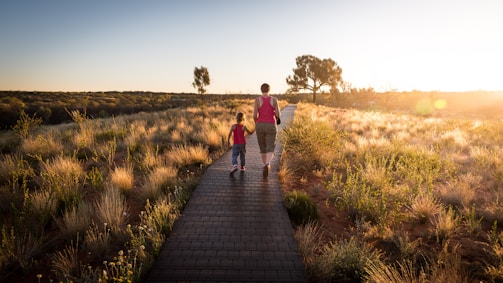 A father and daughter walking hand in hand through a quiet park at sunset.