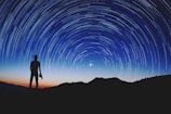Silhouette of a person standing on a hilltop, gazing at the night sky filled with a swirl of star trails. A camera is held in one hand, and the backdrop is a gradient of deep blue and orange from the setting sun.
