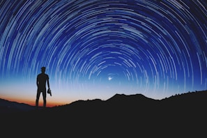 Silhouette of a person standing on a hilltop, gazing at the night sky filled with a swirl of star trails. A camera is held in one hand, and the backdrop is a gradient of deep blue and orange from the setting sun.
