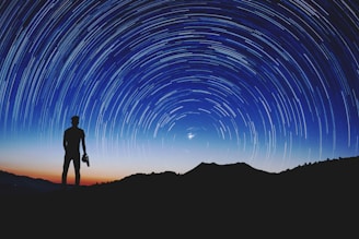Silhouette of a person standing on a hilltop, gazing at the night sky filled with a swirl of star trails. A camera is held in one hand, and the backdrop is a gradient of deep blue and orange from the setting sun.