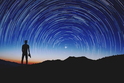 Silhouette of a person standing on a hilltop, gazing at the night sky filled with a swirl of star trails. A camera is held in one hand, and the backdrop is a gradient of deep blue and orange from the setting sun.