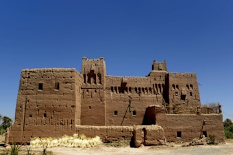 An ancient mud-brick structure with intricate architectural details stands under a clear blue sky. The building exhibits traditional design elements including arched windows and a crenellated roof. The foreground features some straw or dry grass, and surrounding the building are sparse patches of vegetation.
