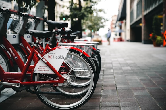 A row of red bicycles is lined up at a bike-sharing station on a city street. The pavement is made of grayish brick tiles, and there are buildings and trees in the background. The bicycles have a white panel with text indicating they are powered by UC Health and are part of Cincinnati's Academic Health System.