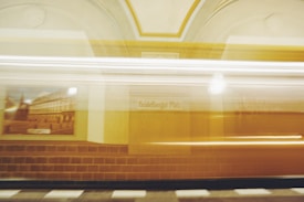 A blurred image of a moving train passing through a station with yellow and beige tones. Signs and architectural details are visible, including a name plate reading 'Heidelberger Platz'. The motion blur conveys a sense of speed.