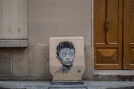 A piece of street art depicting a young boy's face wearing glasses on a building facade. The artwork is on a rectangular canvas leaning against a textured wall between a brown wooden door and a vented panel. The urban setting gives a sense of everyday life and hidden creativity.