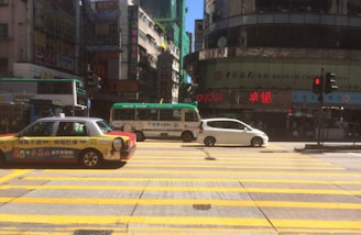 A busy urban street scene with a taxi and other vehicles waiting at a crosswalk. The yellow and black taxi is parked in the foreground, while a green and white minibus is in the middle of the image. Behind them is a white car. There are multiple signs in both English and Chinese characters on tall buildings, including a Bank of China sign. Pedestrians are visible near the buildings, and a traffic light is showing red.