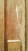 A close-up view of a wooden surface covered in sawdust. A small rectangular piece of wood is placed at the bottom. The texture of the wood grain and the scattered dust particles are visible, with a small wood shaving on the surface.