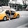 A yellow taxi is parked with its back door open, revealing a person sitting inside with their legs partially outside the vehicle. The taxi has an advertisement for Jay-Z's album '4:44' on top. The background shows a city street with buildings, some under construction, and a few parked cars.