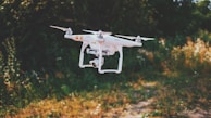 A white drone with four propellers hovers in the air against a blurred natural background of green and brown foliage. The drone has a camera attached, and the setting appears to be an outdoor park or garden.