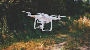 A white drone with four propellers hovers in the air against a blurred natural background of green and brown foliage. The drone has a camera attached, and the setting appears to be an outdoor park or garden.