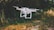 A white drone with four propellers hovers in the air against a blurred natural background of green and brown foliage. The drone has a camera attached, and the setting appears to be an outdoor park or garden.