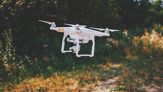 A white drone with four propellers hovers in the air against a blurred natural background of green and brown foliage. The drone has a camera attached, and the setting appears to be an outdoor park or garden.