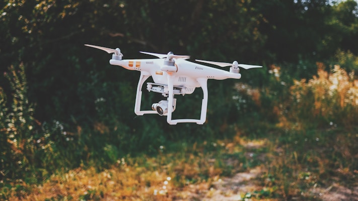 A white drone with four propellers hovers in the air against a blurred natural background of green and brown foliage. The drone has a camera attached, and the setting appears to be an outdoor park or garden.