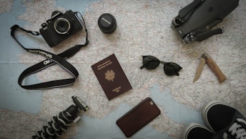 A collection of travel-related items is arranged on a map. Items include a camera with a strap, a lens cap, a passport, sunglasses, a knife, a smartphone, a tripod, and parts of a pair of shoes. All these objects suggest preparation for a journey.