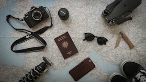 A collection of travel-related items is arranged on a map. Items include a camera with a strap, a lens cap, a passport, sunglasses, a knife, a smartphone, a tripod, and parts of a pair of shoes. All these objects suggest preparation for a journey.