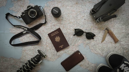 A collection of travel-related items is arranged on a map. Items include a camera with a strap, a lens cap, a passport, sunglasses, a knife, a smartphone, a tripod, and parts of a pair of shoes. All these objects suggest preparation for a journey.
