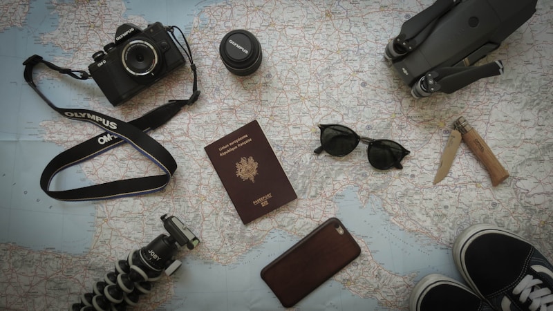 A collection of travel-related items is arranged on a map. Items include a camera with a strap, a lens cap, a passport, sunglasses, a knife, a smartphone, a tripod, and parts of a pair of shoes. All these objects suggest preparation for a journey.