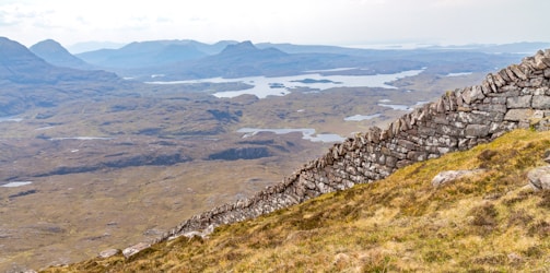 A landscape view featuring a dry stone wall stretching along a hillside. The wall is constructed with large, uneven stones that create a rugged appearance. The background consists of a vast expanse of rolling hills, a patchwork of marshy lands, and numerous small lakes. Distant mountain ranges form a hazy backdrop, creating a sense of depth and vastness in the scenery.