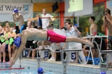 A swimmer in vibrant red swim trunks and a blue swim cap is diving into a pool from a starting block labeled '3'. Several people, including competitors and spectators, are gathered around at the poolside, with some wearing colorful swimsuits.