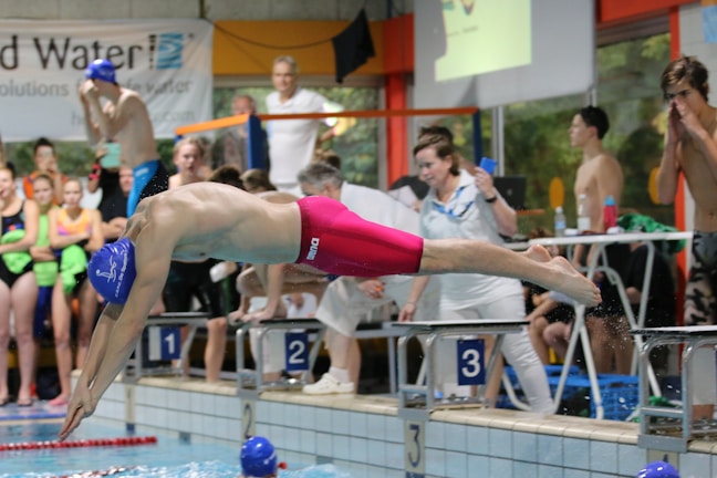 A swimmer in vibrant red swim trunks and a blue swim cap is diving into a pool from a starting block labeled '3'. Several people, including competitors and spectators, are gathered around at the poolside, with some wearing colorful swimsuits.