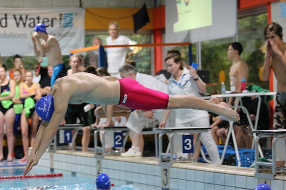 A swimmer in vibrant red swim trunks and a blue swim cap is diving into a pool from a starting block labeled '3'. Several people, including competitors and spectators, are gathered around at the poolside, with some wearing colorful swimsuits.