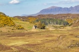 A rustic stone house is nestled in a lush, green valley surrounded by hills covered with yellow-flowered shrubs. In the background, expansive mountains rise under a partly cloudy sky. The landscape is dotted with trees, and the area appears remote and tranquil.