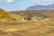 A rustic stone house is nestled in a lush, green valley surrounded by hills covered with yellow-flowered shrubs. In the background, expansive mountains rise under a partly cloudy sky. The landscape is dotted with trees, and the area appears remote and tranquil.