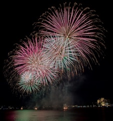 Bright and colorful fireworks burst in the night sky above a calm body of water. The fireworks display features prominent pink and green hues radiating outward, illuminating the surrounding area. In the background, city lights are visible along the shoreline, adding to the festive atmosphere.