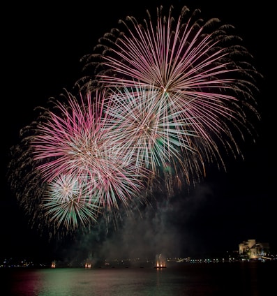 Bright and colorful fireworks burst in the night sky above a calm body of water. The fireworks display features prominent pink and green hues radiating outward, illuminating the surrounding area. In the background, city lights are visible along the shoreline, adding to the festive atmosphere.