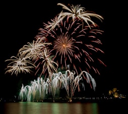 A breathtaking display of fireworks bursts across the night sky above a calm body of water. The bright, colorful explosions create intricate patterns, illuminating the surroundings with hues of red, green, and gold. Reflections of the fireworks dance on the surface of the water, adding to the visual spectacle.