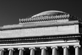 A grand architectural structure with classical columns and ornate detailing, featuring inscriptions on the facade, including references to a college founded in New York.