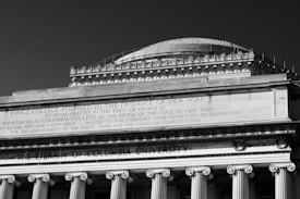A grand architectural structure with classical columns and ornate detailing, featuring inscriptions on the facade, including references to a college founded in New York.
