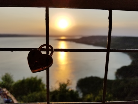 A heart-shaped lock is attached to a fence, set against the backdrop of a serene sunset over a large body of water. The sun is low, casting a warm glow and reflecting on the water surface. Lush greenery can be seen in the foreground, while distant hills create a silhouette on the horizon.